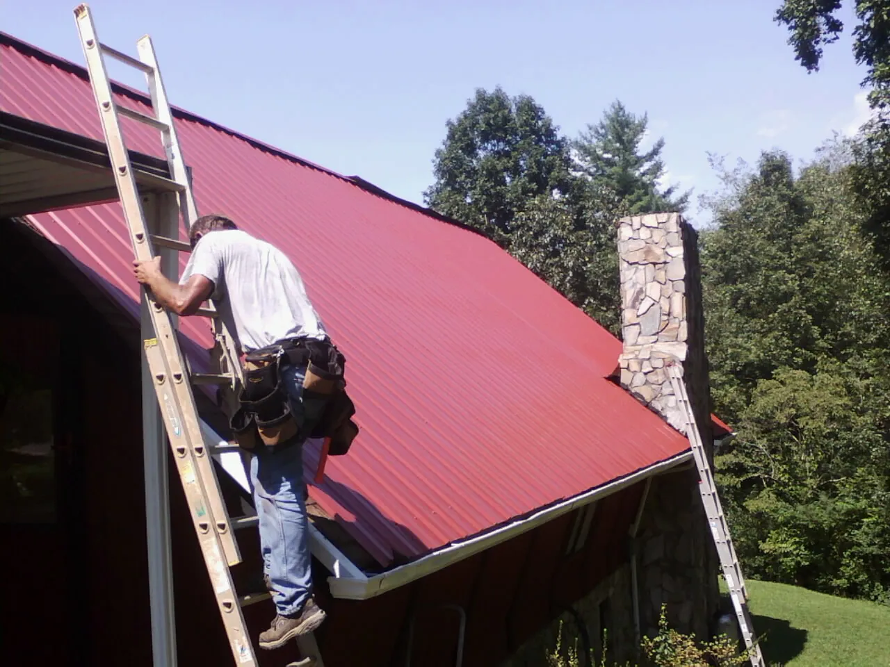 Roofer on a ladder beside a red metal roof and stone chimney — Soffit & Fascia Repair by Roofing Brothers in Dobbs Ferry